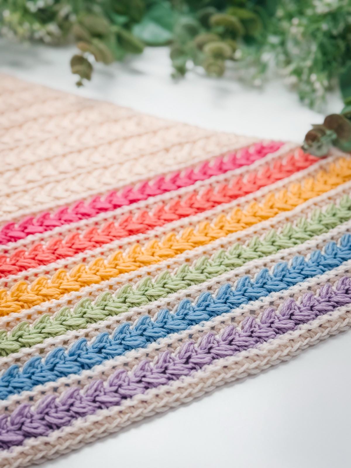 A crocheted fabric with colorful horizontal stripes in purple, blue, green, yellow, orange, and pink rests on a white surface with green foliage in the background—perfect as a handmade gift.
