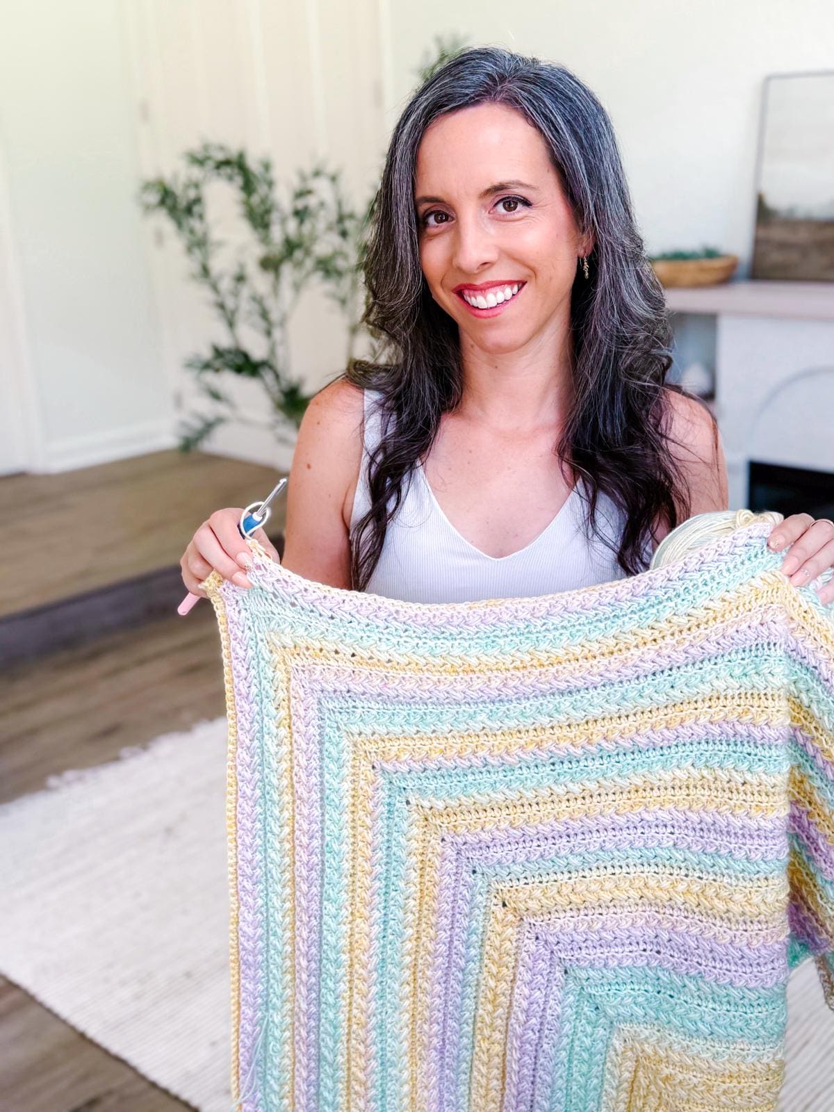 A woman with long brown hair wearing a colorful crocheted shawl and unique jewellery stands in a bright room with a white rug and potted plant.