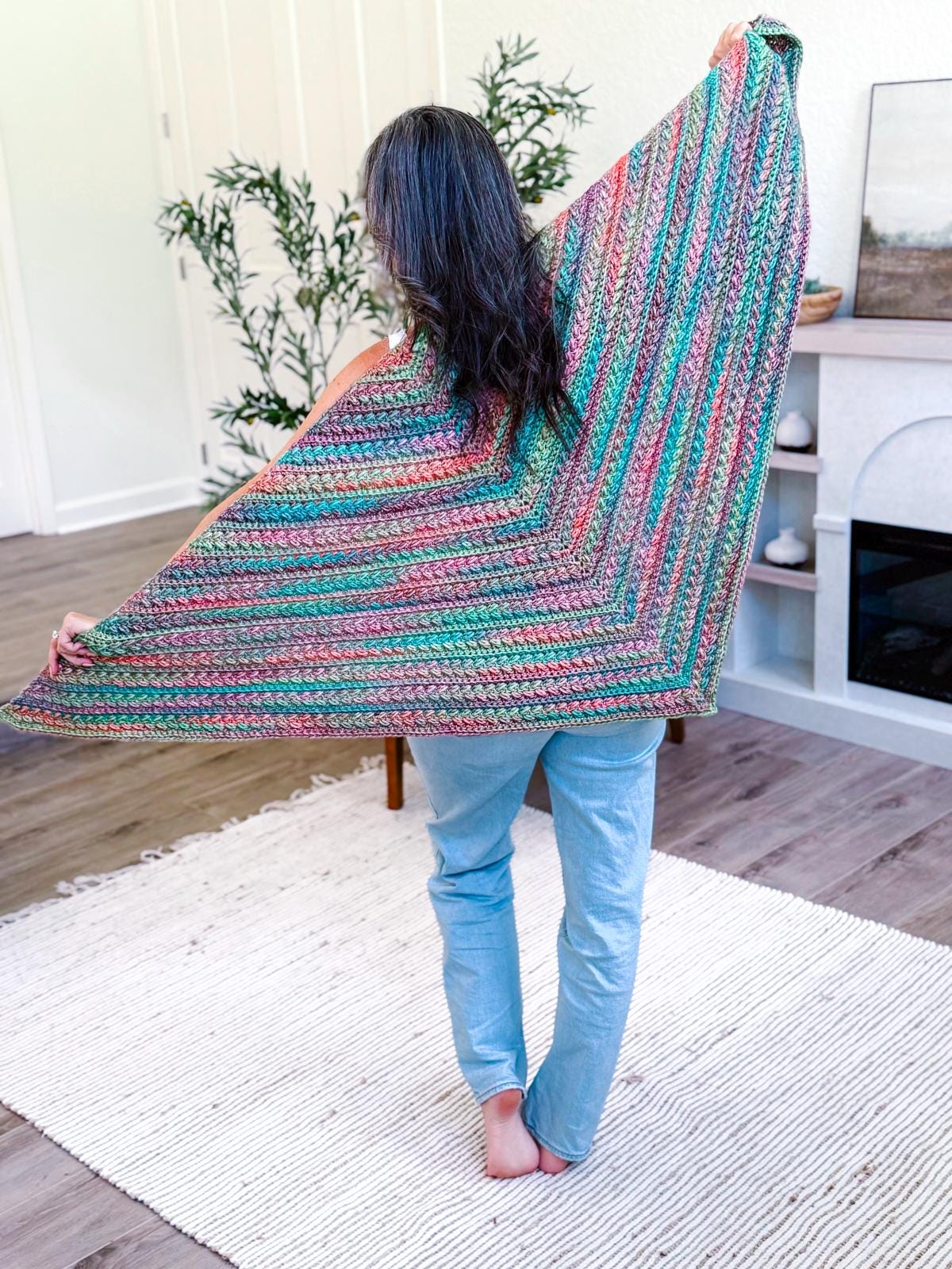 Person standing barefoot on a rug, holding out a large, multicolored knit shawl—a perfect handmade gift—in a bright room with plants and modern decor.