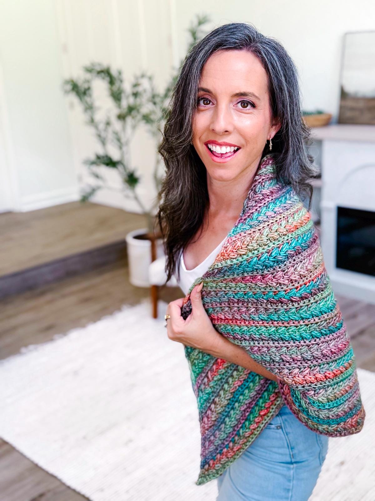 A woman with long brown hair wearing a colorful crocheted shawl and unique jewellery stands in a bright room with a white rug and potted plant.
