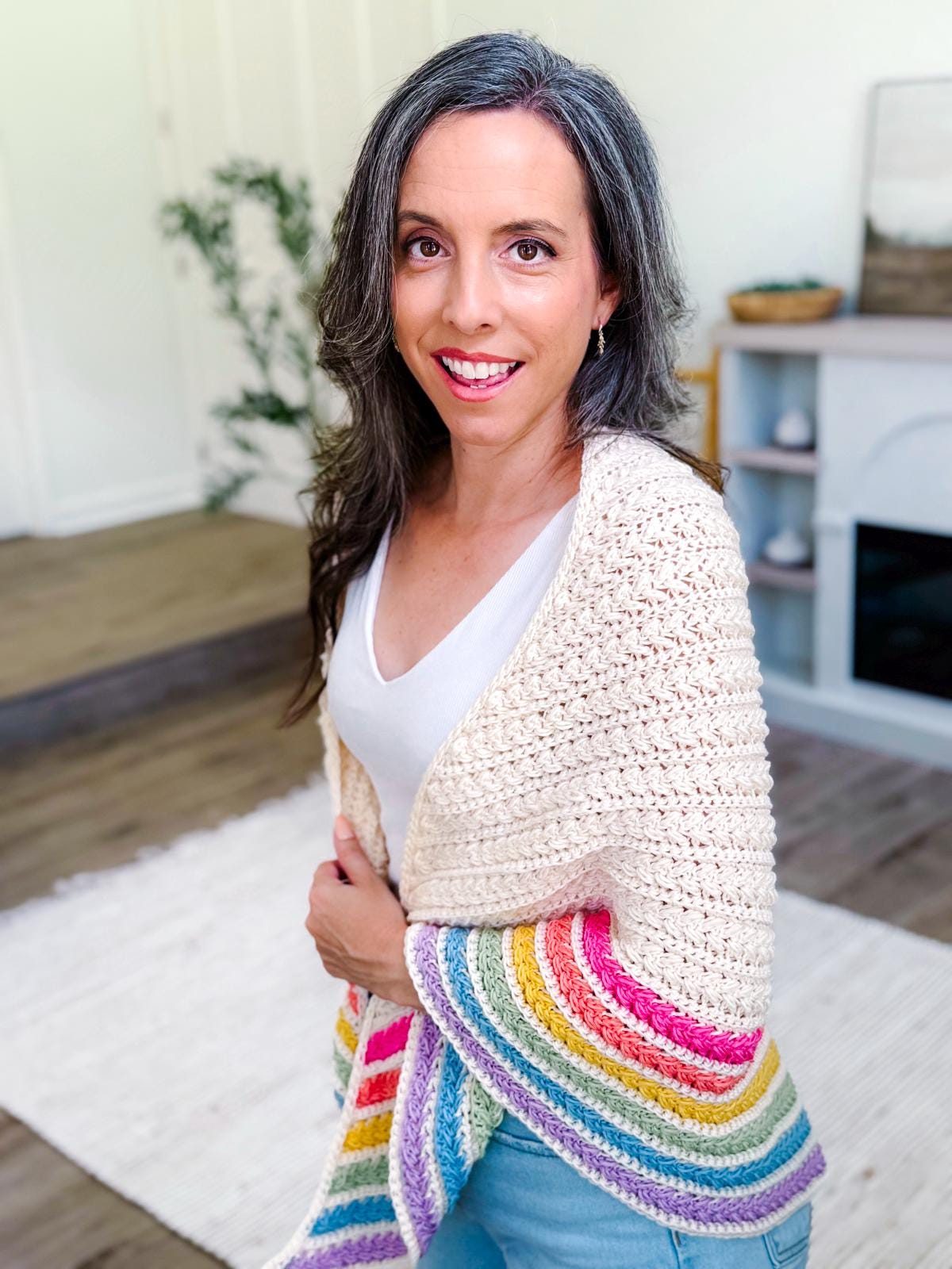 A woman with long brown hair and gray streaks smiles while wearing a cream-colored crochet shawl with rainbow stripes on the edge, standing in a bright, modern room—her look completed by unique jewellery.
