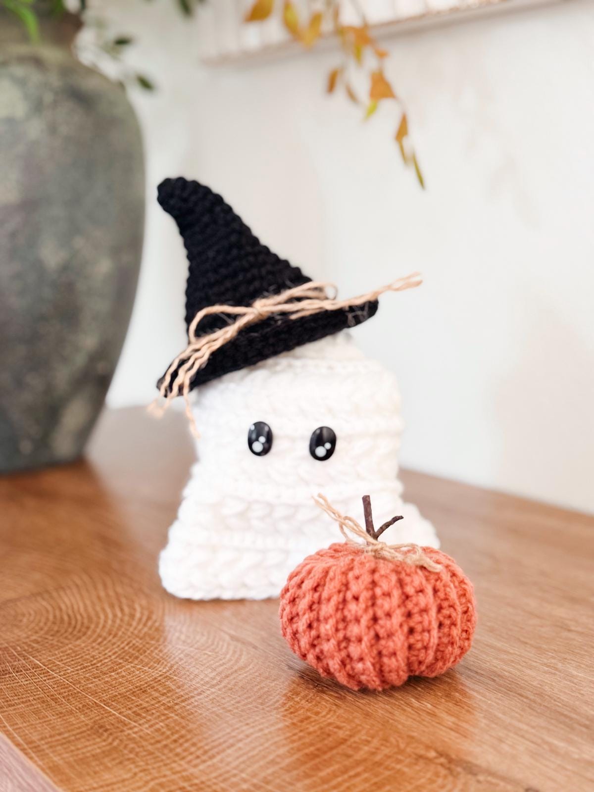 A crocheted ghost with a black witch hat and string bow sits on a wooden surface next to a small crocheted orange pumpkin, making a charming Halloween Crochet Home Decor.