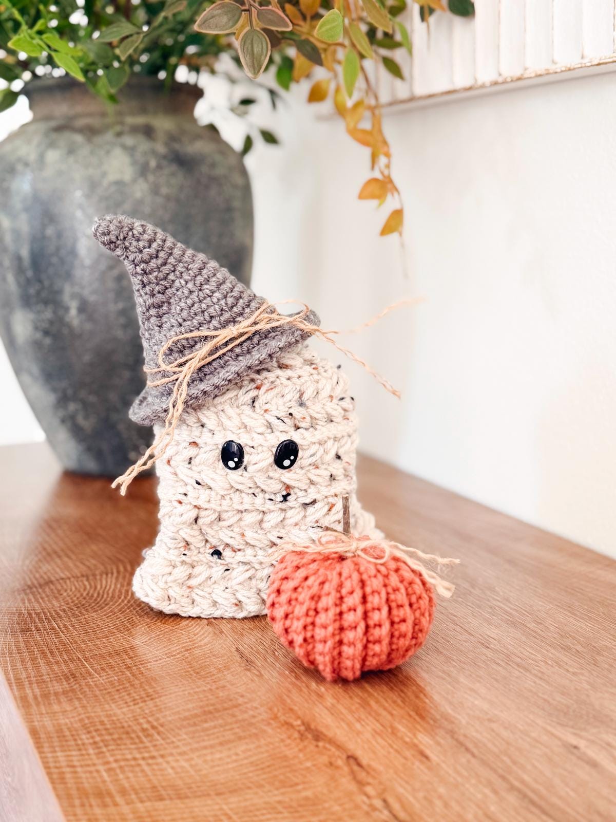 A crocheted ghost with a gray witch hat and raffia bow sits next to a small orange crocheted pumpkin on a wooden surface, making a charming handmade gift for lovers of whimsical decor.