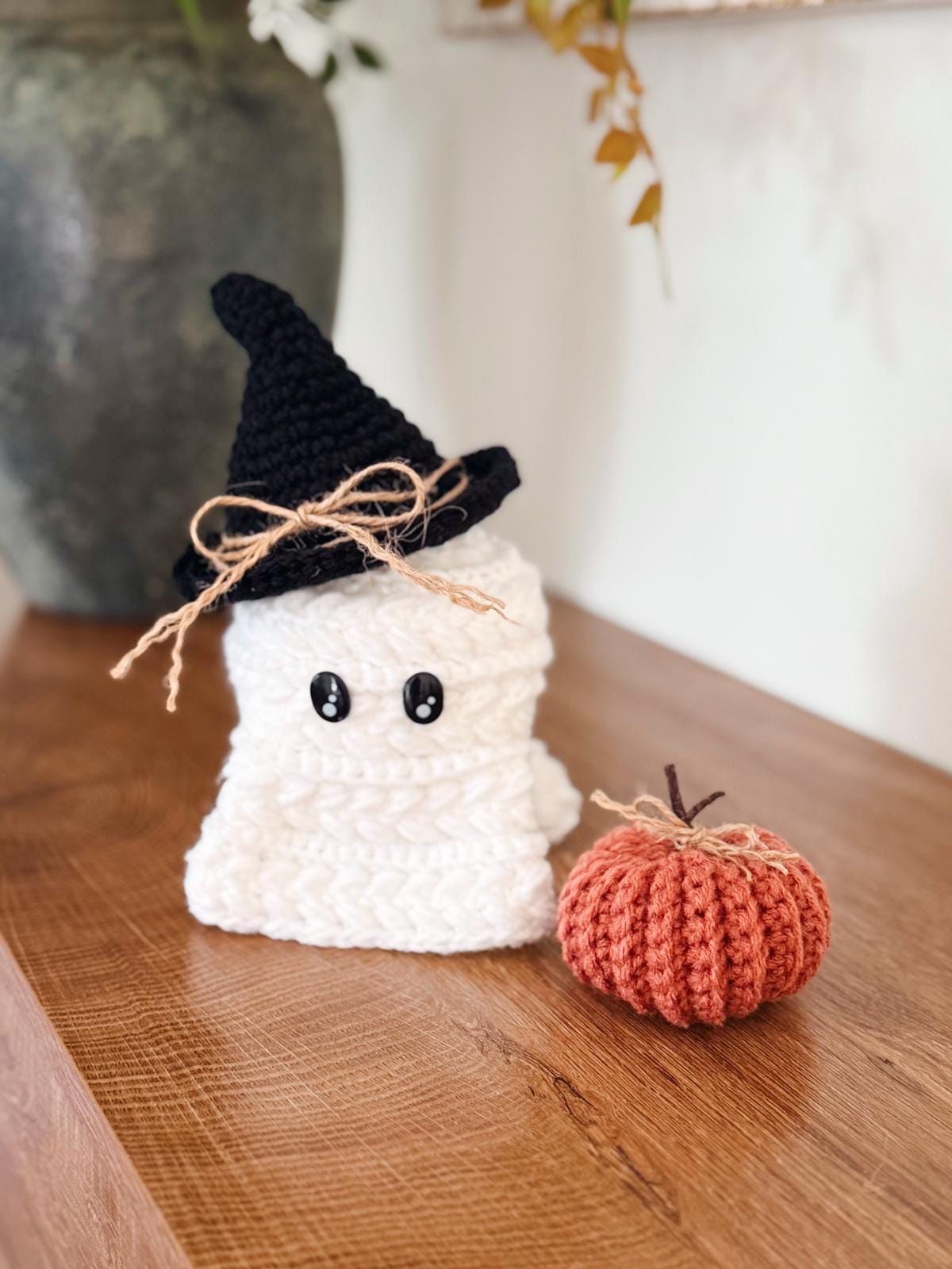 A crocheted ghost with a black witch hat and twine bow sits on a wooden surface next to a small crocheted pumpkin—a charming handmade gift perfect for autumn.