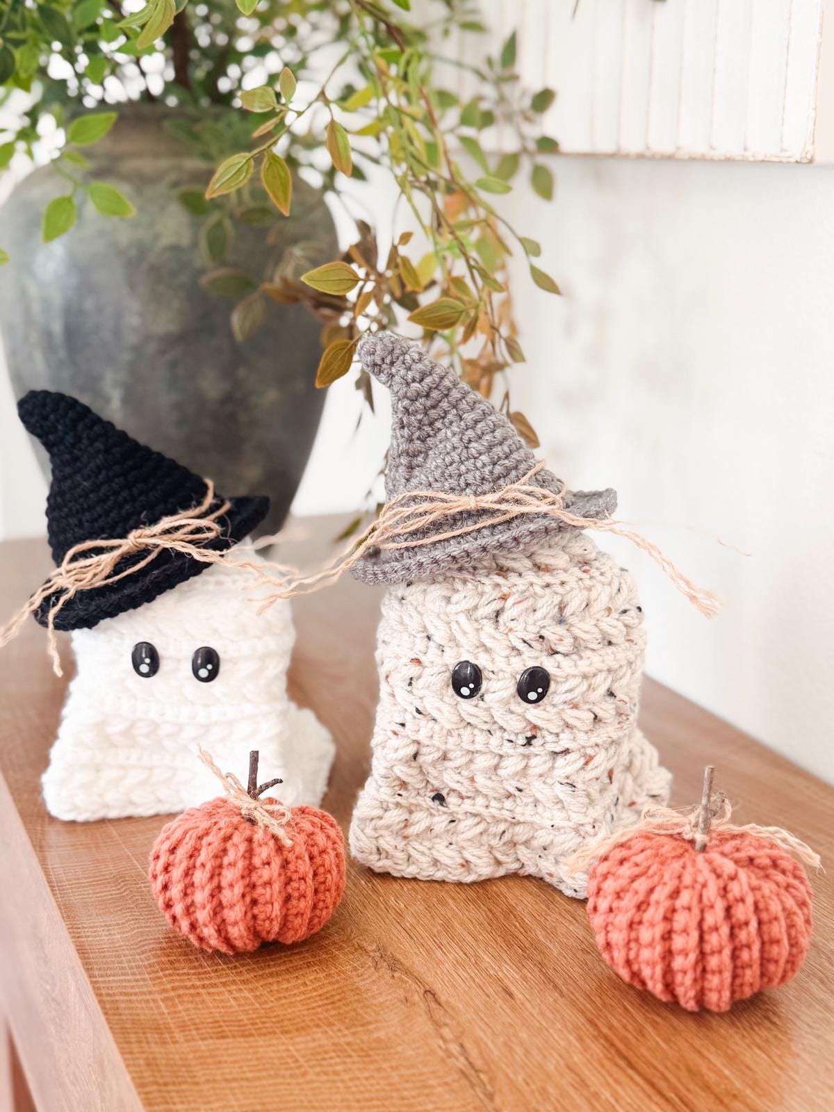 Two crocheted ghost decorations with hats and two crocheted pumpkins are displayed on a wooden surface with a leafy plant in the background—perfect as a handmade gift for Halloween Decor.