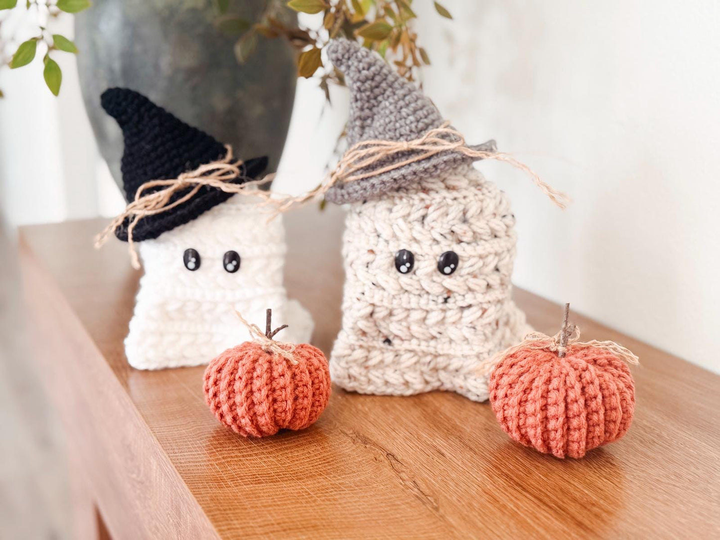 Two crocheted ghost figures wearing hats sit on a wooden surface next to two crocheted pumpkins, making a charming handmade gift. Autumn leaves add a cozy touch in the background.