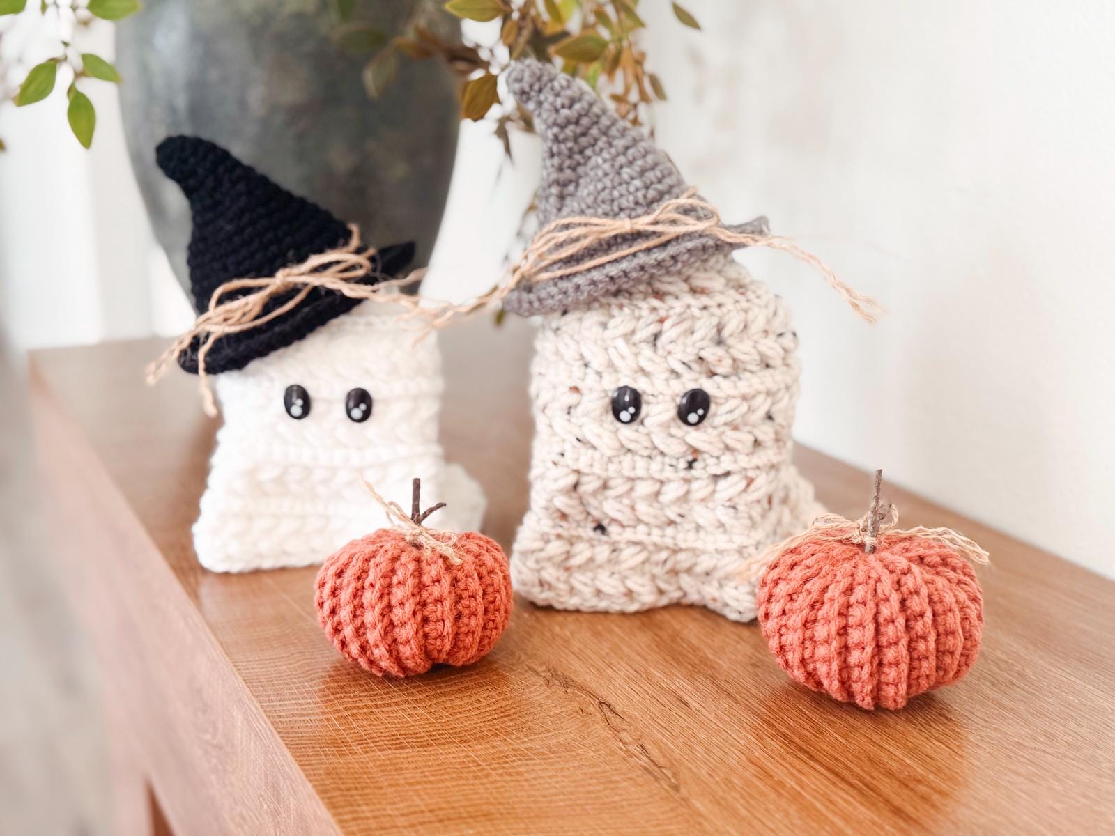 Two crocheted ghost figures wearing hats sit on a wooden surface next to two crocheted pumpkins, making a charming handmade gift. Autumn leaves add a cozy touch in the background.