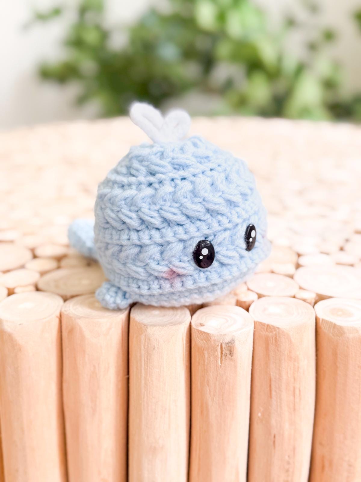A small, crocheted blue whale plush with black eyes sits on a light wooden surface—perfect as a charming handmade gift from an easy crochet pattern.