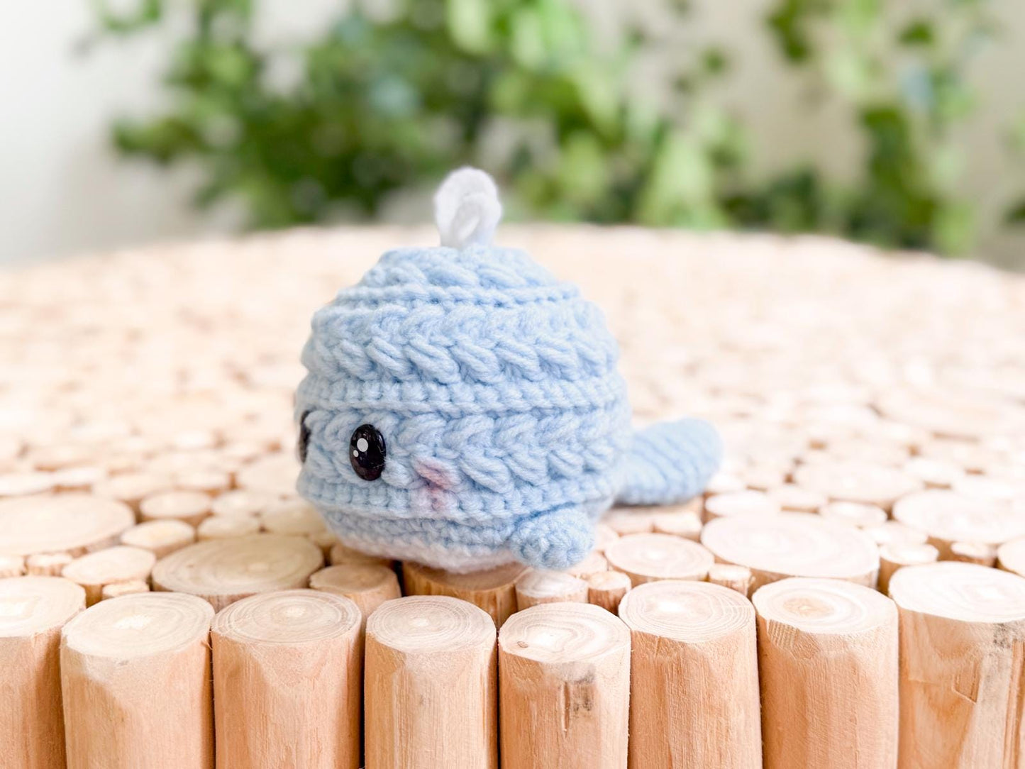 A small, crocheted blue whale plush with black eyes sits on a light wooden surface—perfect as a charming handmade gift from an easy crochet pattern.