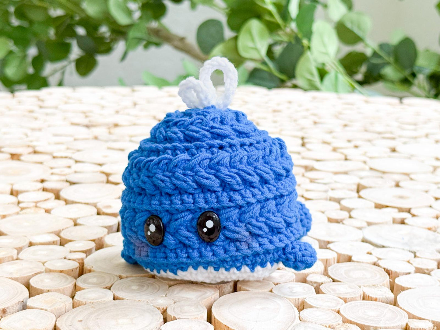 A small, crocheted blue whale with large black eyes and a white spout sits on a wooden surface with green leaves in the background, making a charming handmade gift.