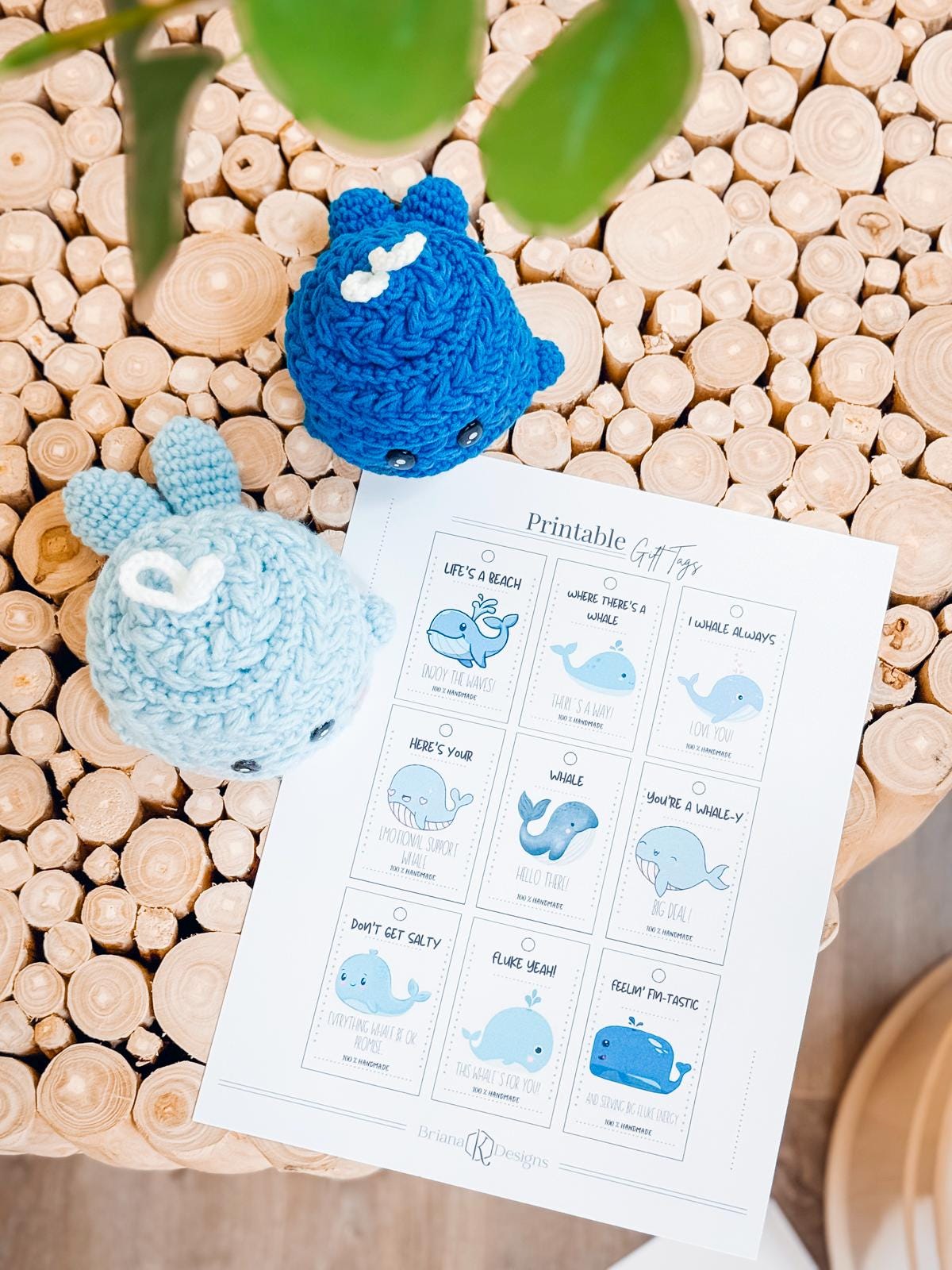 Two crocheted whale toys and a sheet of printable gift tags with whale designs rest on a round wooden table, perfect as a charming handmade gift.