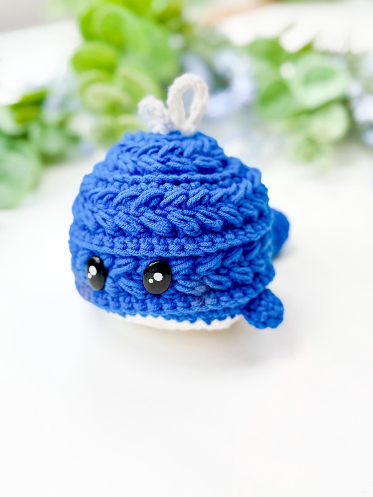 A small, crocheted blue whale plush with black eyes and white fins sits on a white surface, with green foliage in the blurred background—perfect as a charming handmade gift.