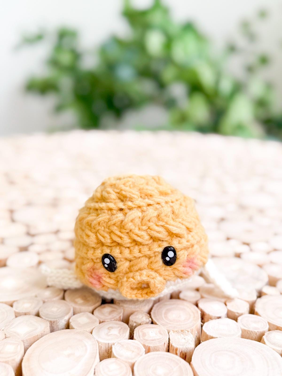 A small, crocheted yellow plush fish toy with black eyes and pink cheeks sits on a textured wooden surface, making a charming handmade gift. A leafy green plant in the background adds to its cozy appeal.