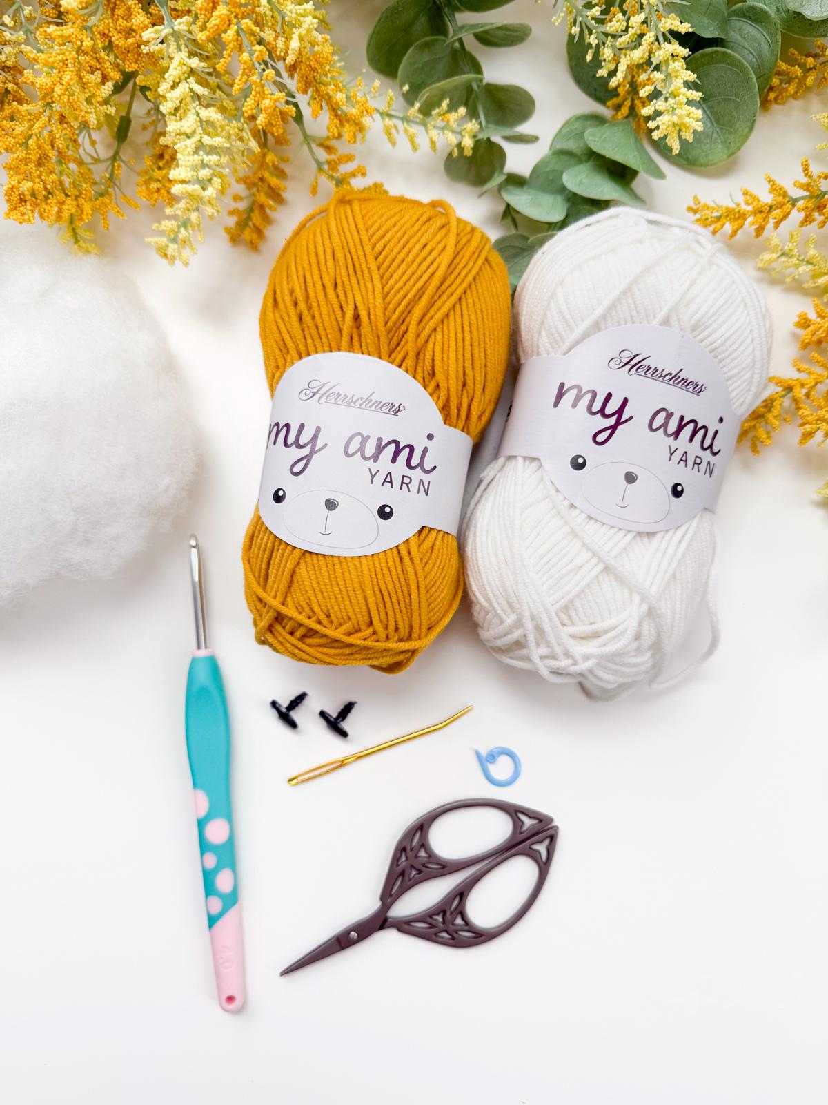 Two skeins of yellow and white yarn, polyester stuffing, crochet hook, scissors, safety eyes, yarn needle, and stitch marker arranged on a white surface with flowers—perfect supplies for crafting a charming handmade gift.