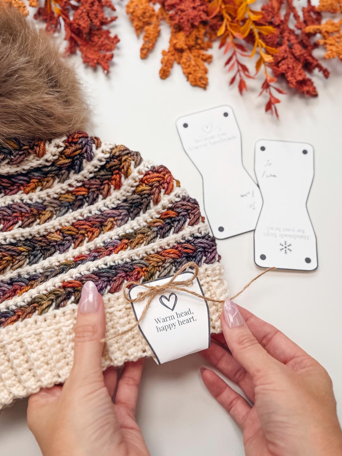 A person ties a tag reading "Warm head, happy heart." onto a knitted hat, with crocheted patterns, autumn leaves, and hints of vintage clothes in the background.