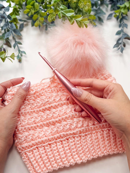 Peach crochet beanie with wheat texture and pink pom pom held in hands with a pink crochet hook, greenery behind