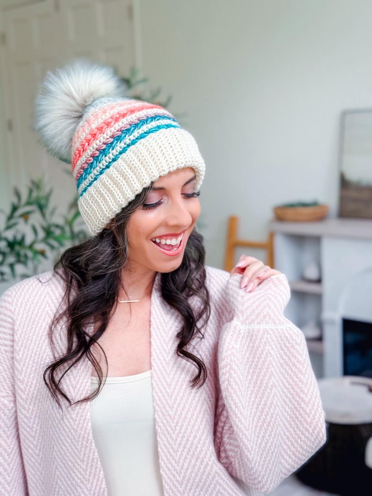 A woman smiles while wearing a crochet hat with colorful stripes and a pom-pom, paired with a textured, light pink cardigan and unique jewellery in a bright indoor setting.