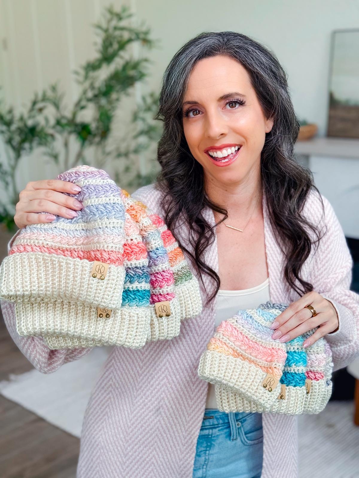 A woman with long dark hair and gray streaks is holding several crochet pastel-colored hats, smiling at the camera indoors, showcasing the perfect handmade gift.