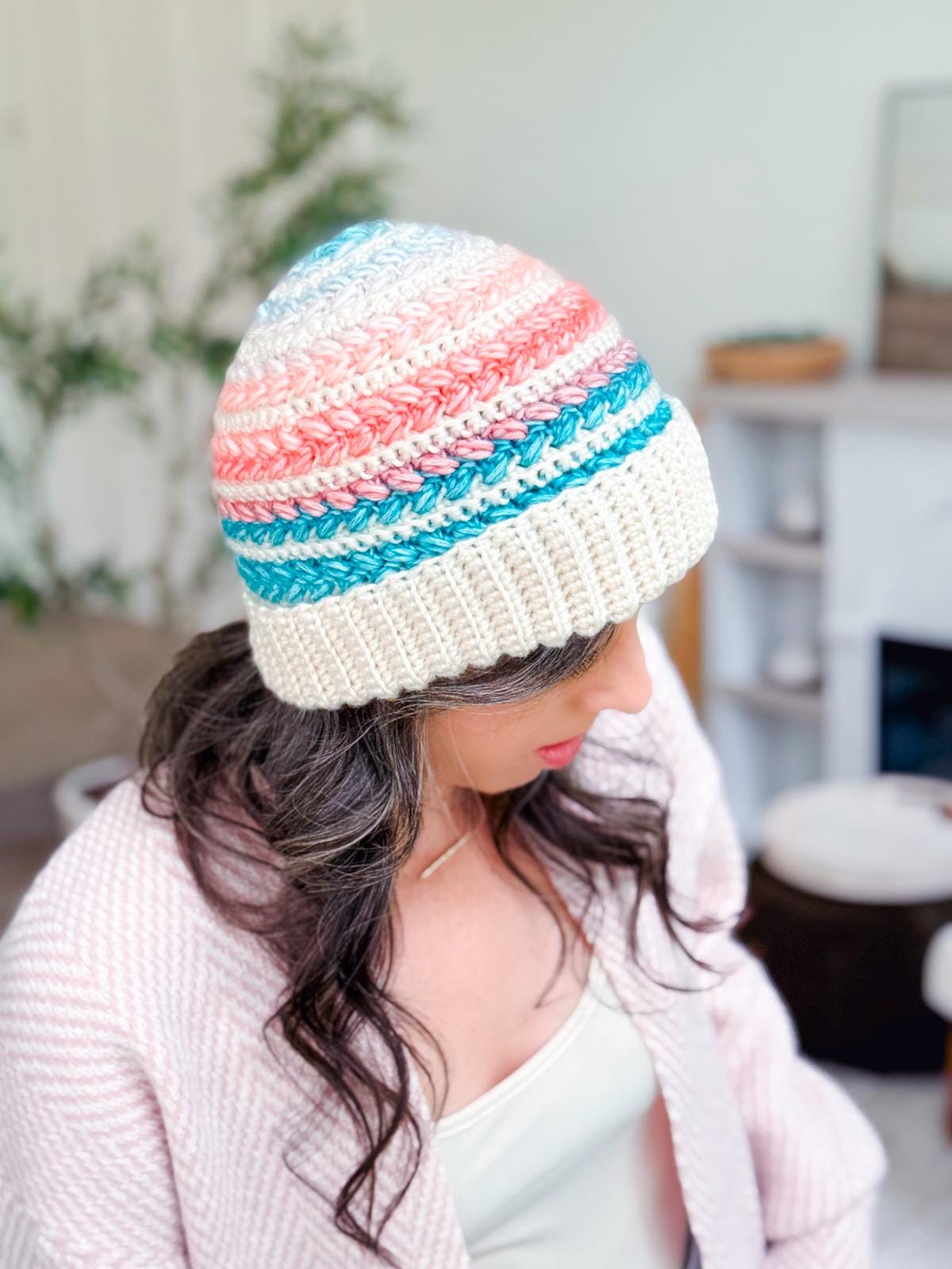 A woman wearing a pastel-colored crocheted beanie and a light pink sweater, paired with unique jewellery, looks down in a bright, cozy room.