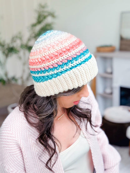 Pastel striped crochet beanie with wide cream brim worn by a woman looking down in a pink cardigan