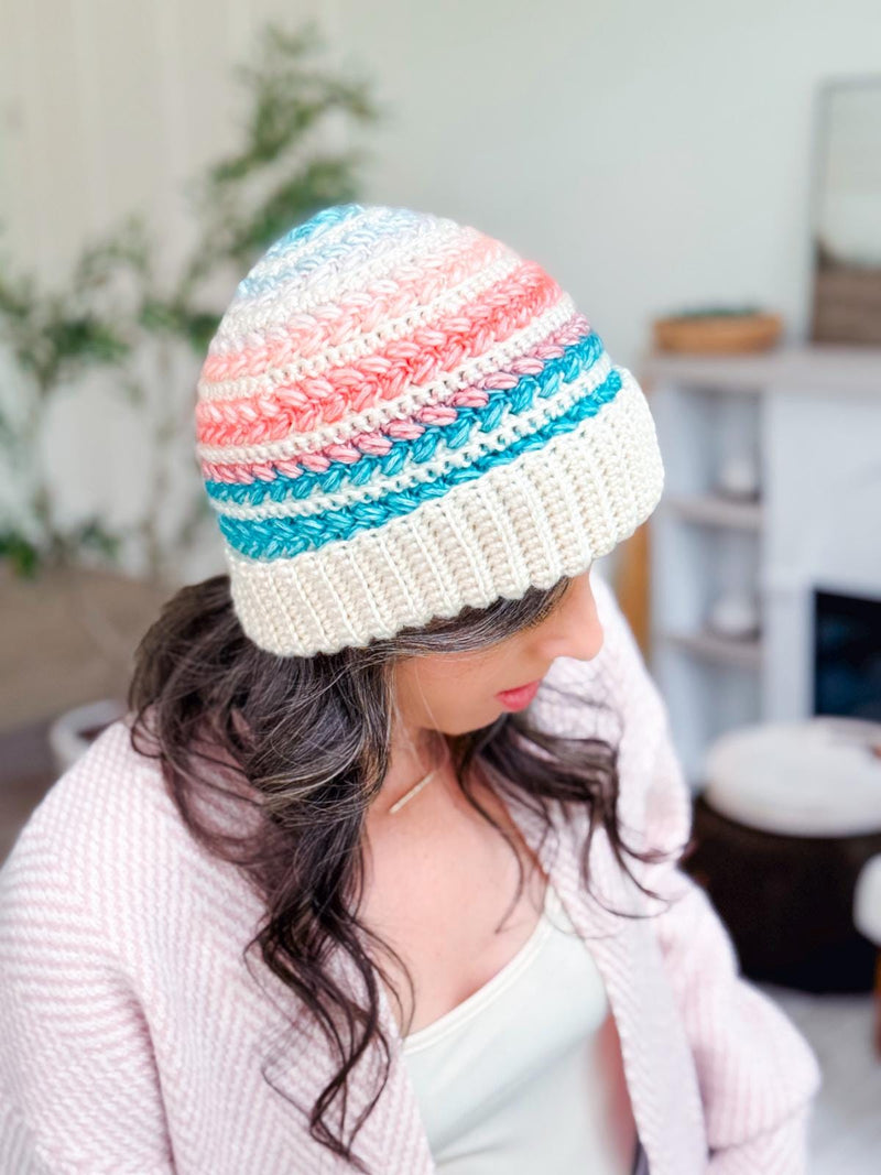 Pastel striped crochet beanie with wide cream brim worn by a woman looking down in a pink cardigan