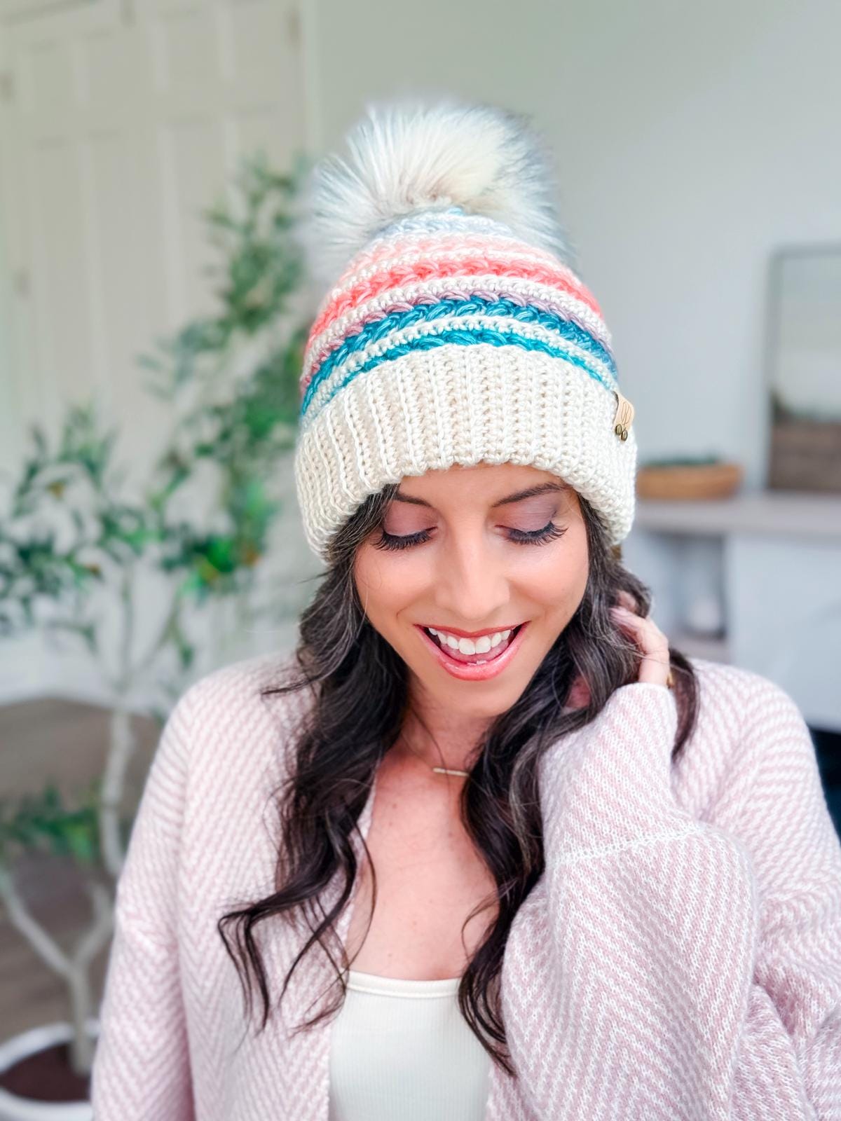 Woman wearing a cream crochet hat with colorful stripes and a pom-pom, a pale pink cardigan, and a white top, smiling indoors—her look finished with unique jewellery for that extra touch.