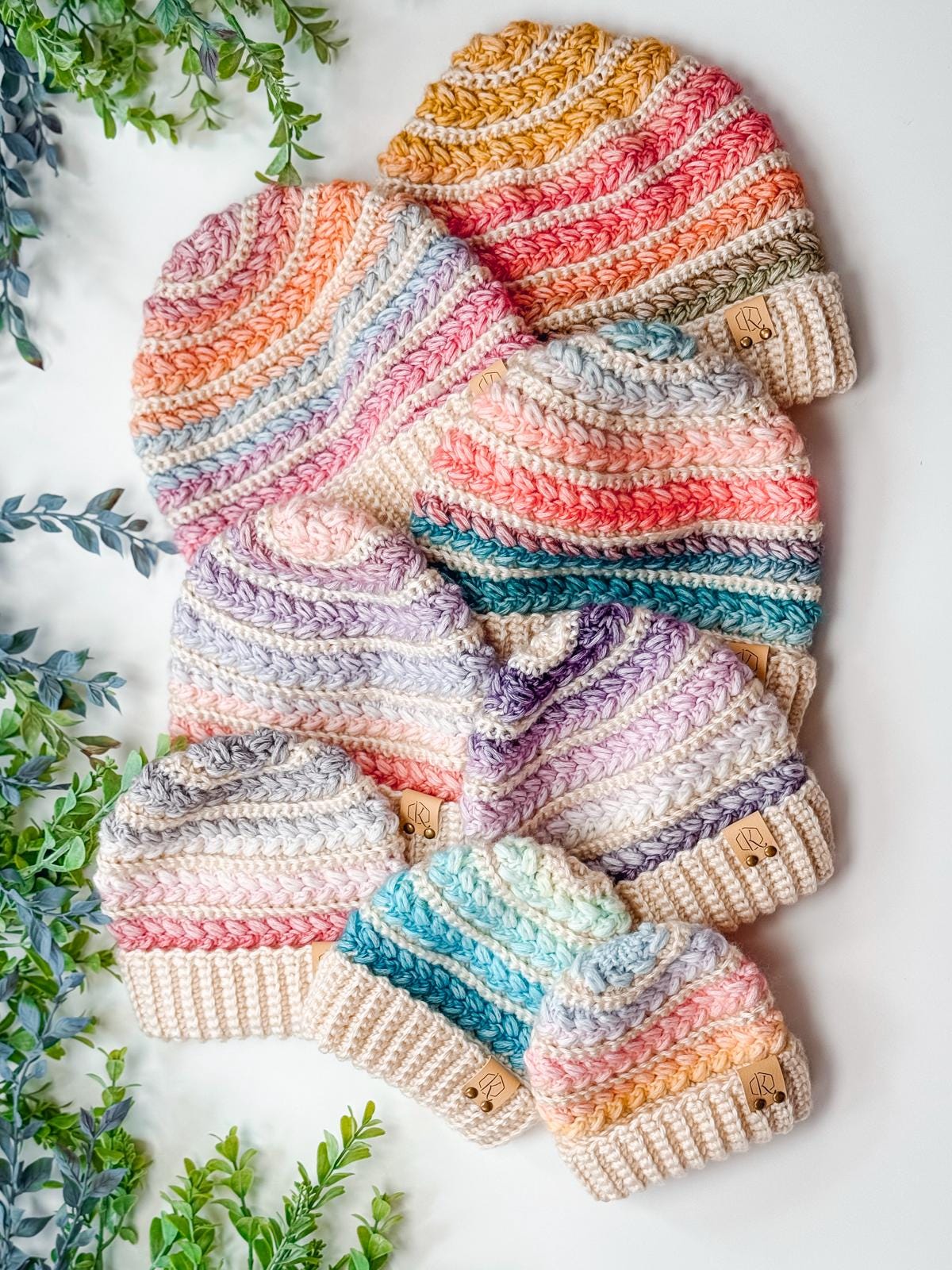 Seven colorful crocheted beanies with striped patterns are arranged on a white surface next to green leafy plants, making a charming handmade gift that pairs perfectly with vintage clothes.