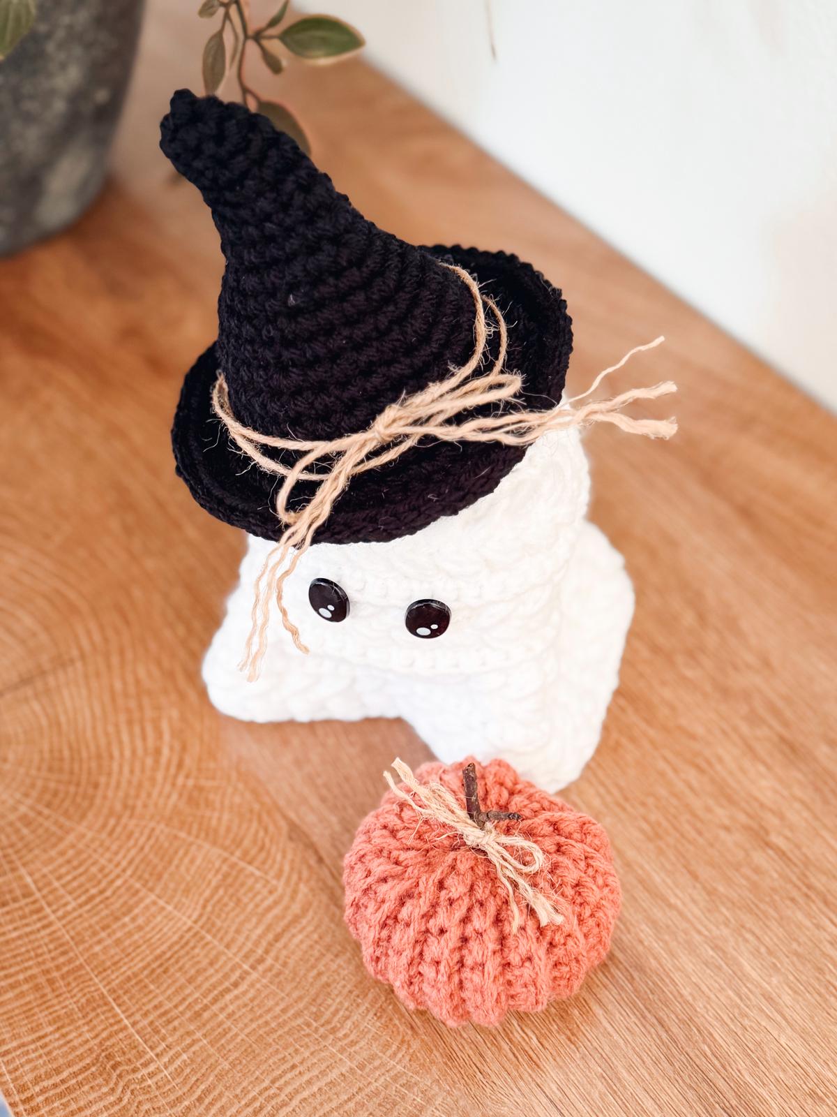 Two crocheted ghost figures wearing hats sit on a wooden surface next to two crocheted pumpkins, making a spooky halloween crochet home decor.