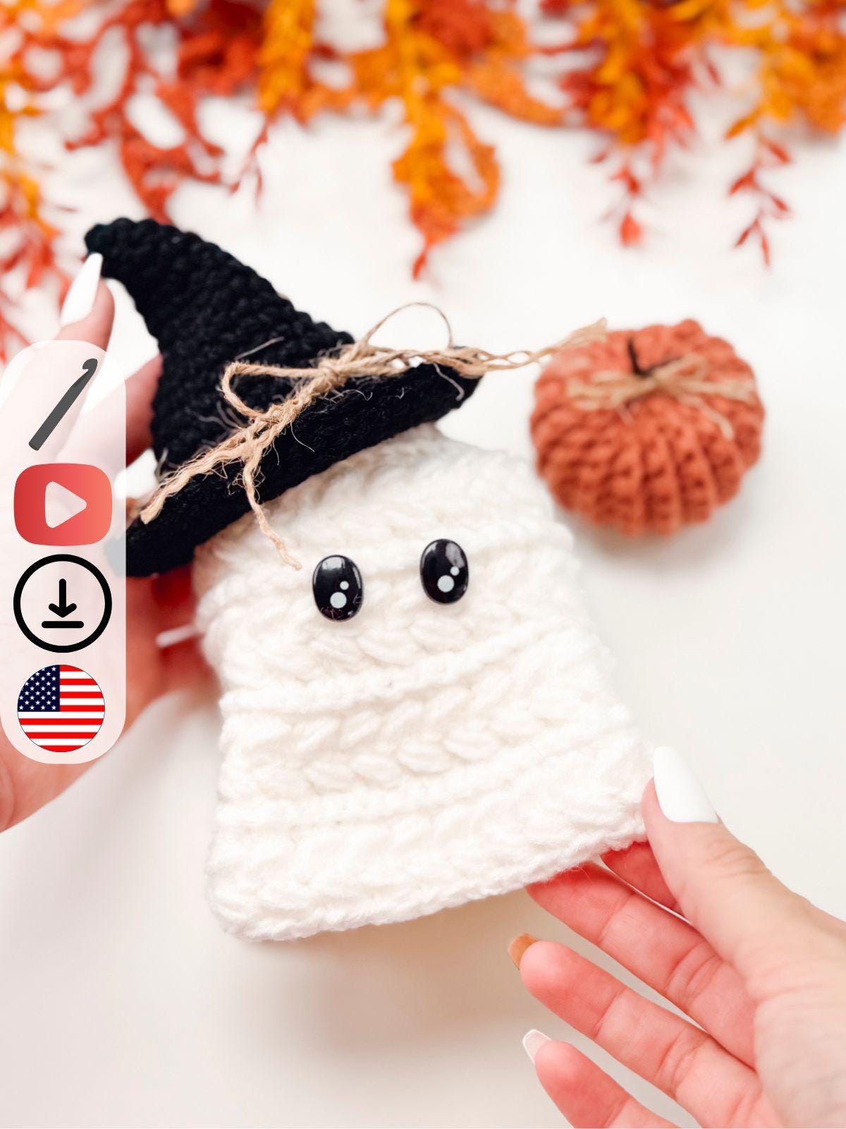 A hand holds a crocheted white ghost with a black witch hat and twine bow—an adorable handmade gift. An orange crocheted pumpkin and autumn leaves are in the background, adding cozy charm.