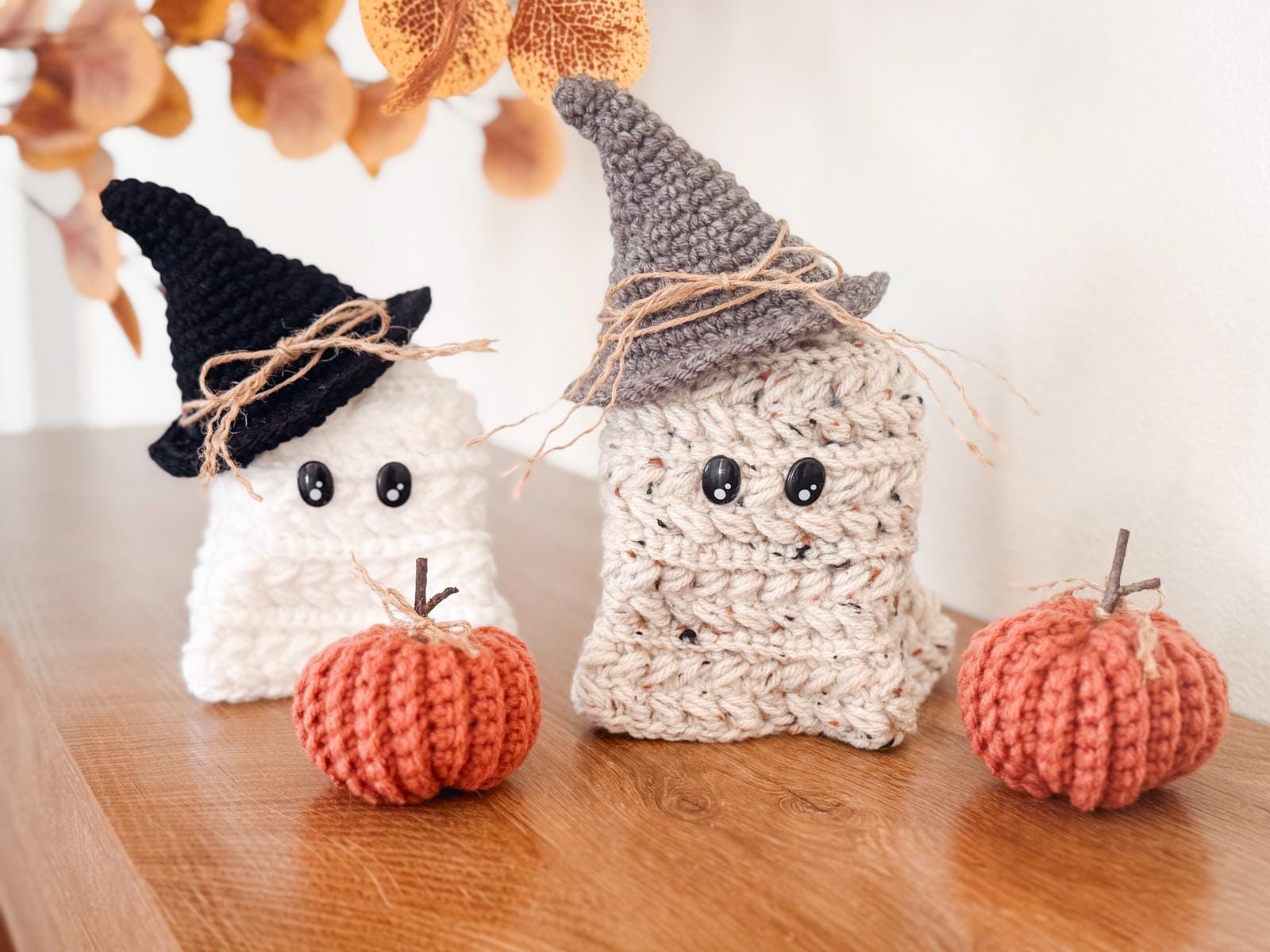 Two crocheted ghost figures wearing hats sit on a wooden surface next to two crocheted pumpkins, making a charming handmade gift. Autumn leaves add a cozy touch in the background.