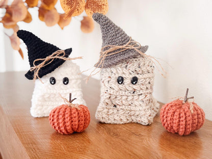 Two crocheted ghost figures wearing hats sit on a wooden surface next to two crocheted pumpkins, making a charming handmade gift. Autumn leaves add a cozy touch in the background.