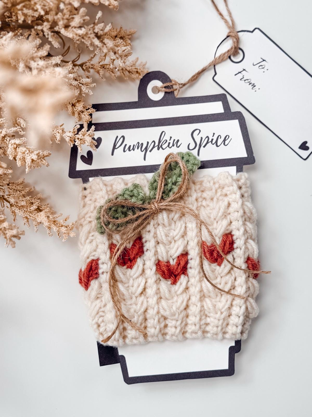 A crocheted cream-colored cozy with red and green accents, tied with twine, displayed on a "Pumpkin Spice" tag with a blank gift tag nearby. Perfect as a handmade gift, with dried foliage in the corner adding charm.