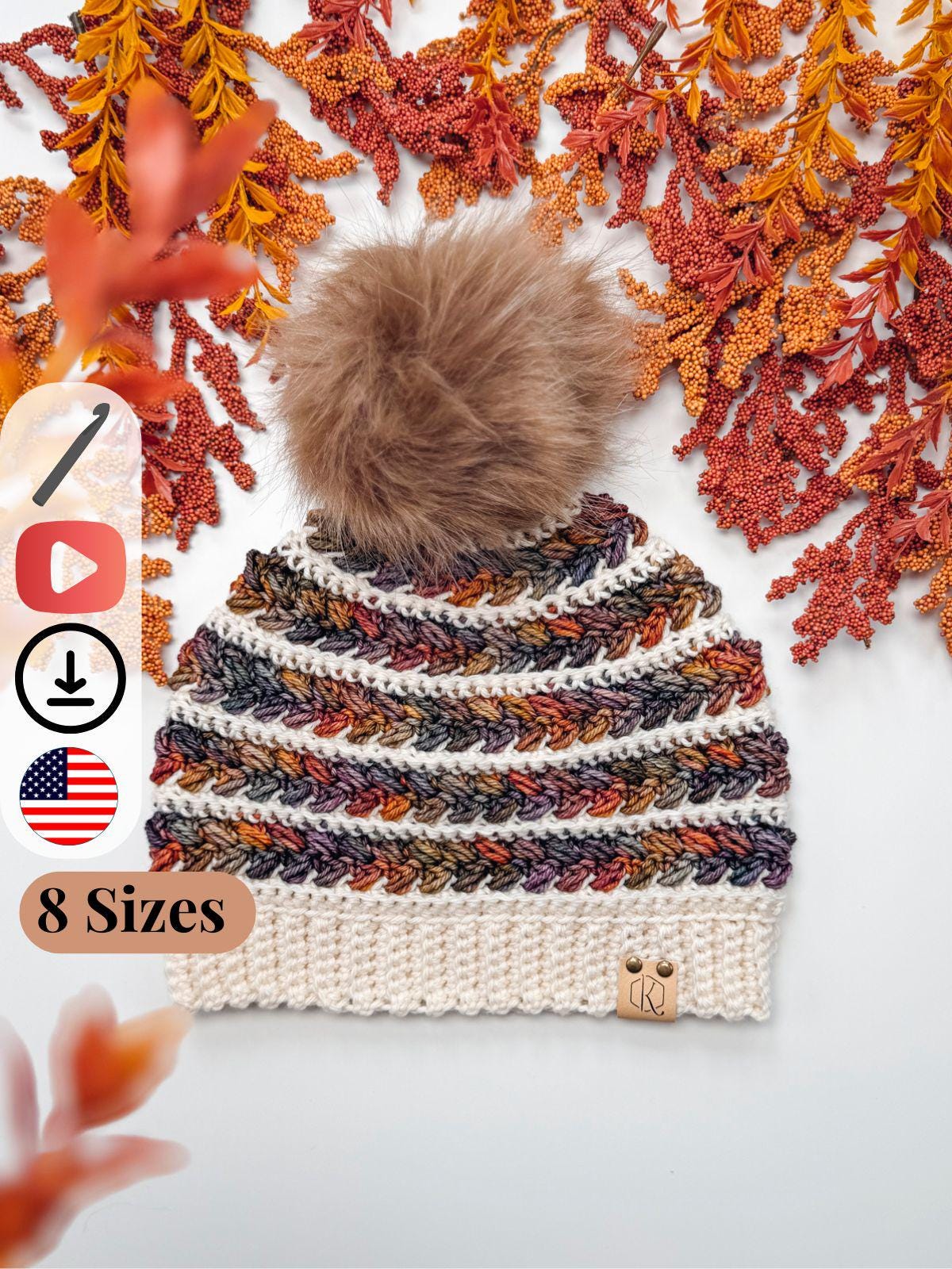 A crocheted hat with multicolored stripes, a white brim, and a large brown pom-pom is displayed on a white surface with orange and yellow foliage, perfect as a cozy handmade gift. Text indicates "8 Sizes.”