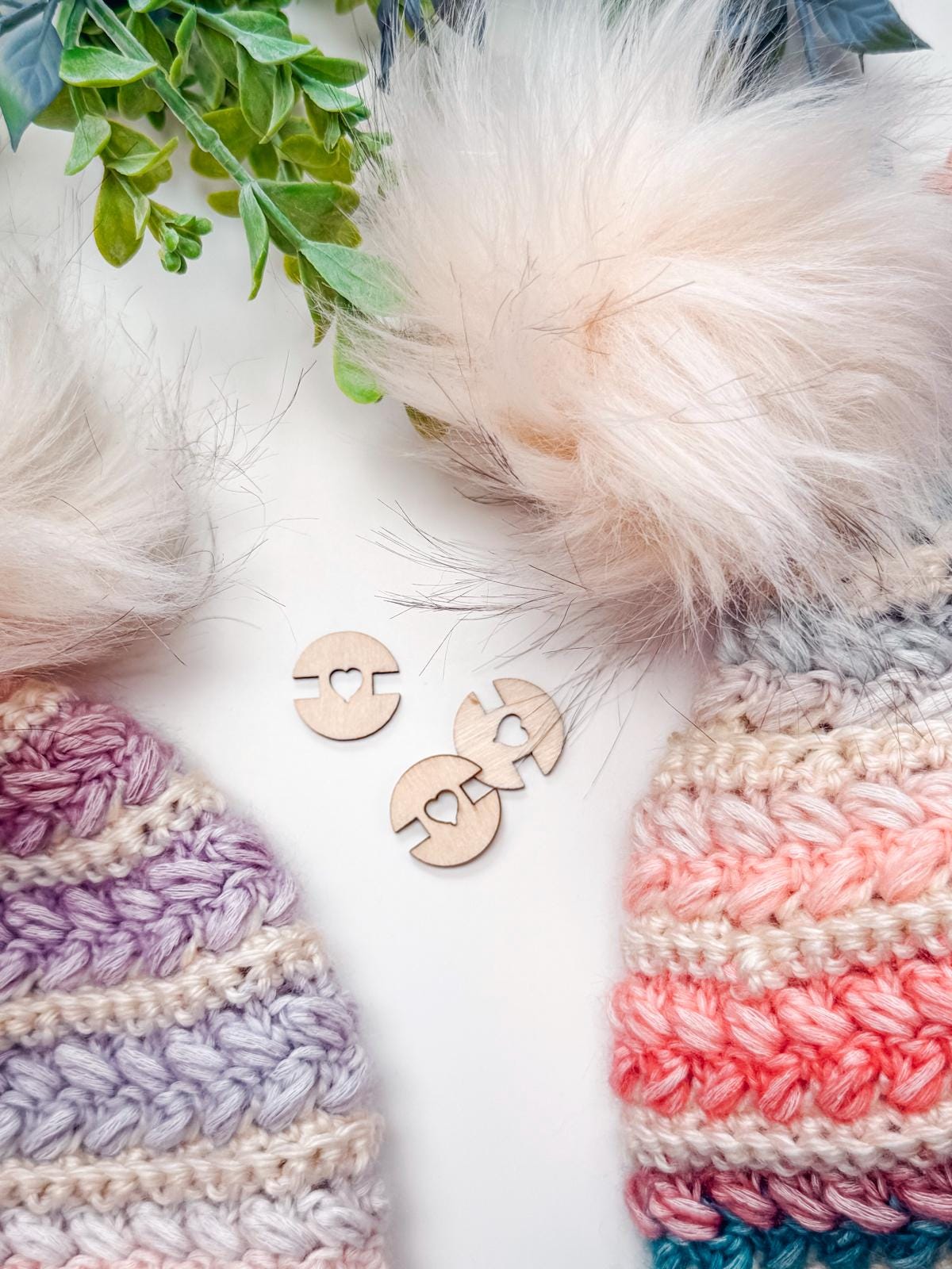 Two crocheted hats with faux fur pom-poms, one in purple tones and one in pink tones, are displayed with three wooden buttons featuring heart cutouts. Green leaves frame the top—an ideal handmade gift for any occasion.