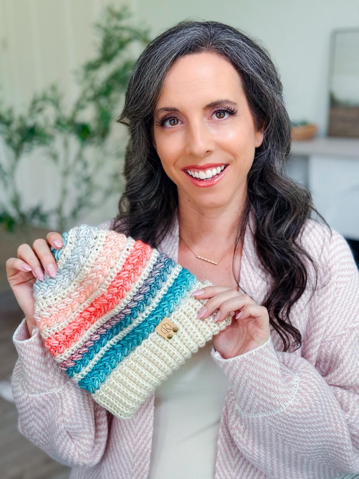 A woman with long, wavy dark hair and a light pink sweater holds up a crochet hat with colorful stripes—a perfect handmade gift. Indoors, with plants and a blurred background.
