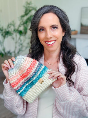 Pink, coral and teal striped crochet beanie held up by a smiling woman with long dark hair in a pink cardigan