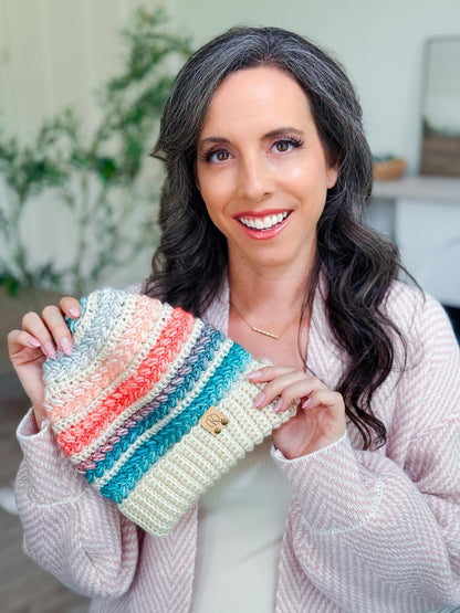 Pink, coral and teal striped crochet beanie held up by a smiling woman with long dark hair in a pink cardigan