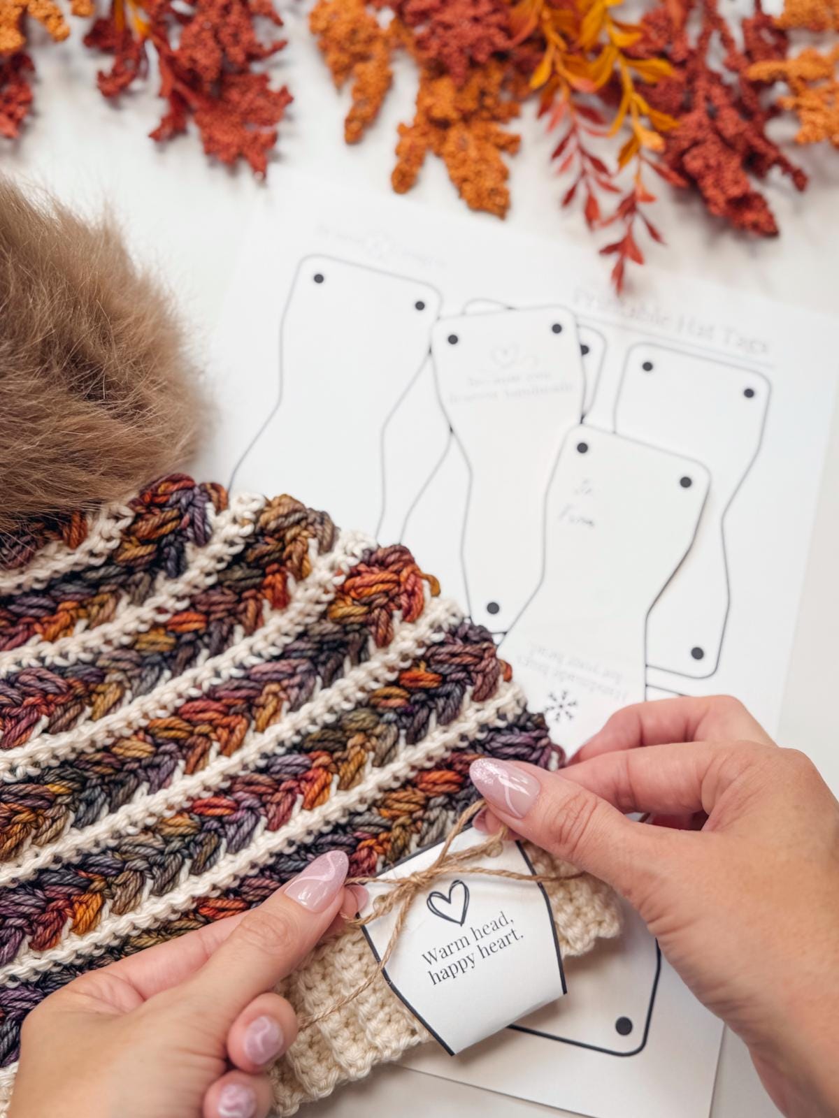 A person attaches a label reading "Warm heart, happy heart" to a crochet hat with autumn-colored yarn; handmade gift tag templates and orange foliage are in the background.