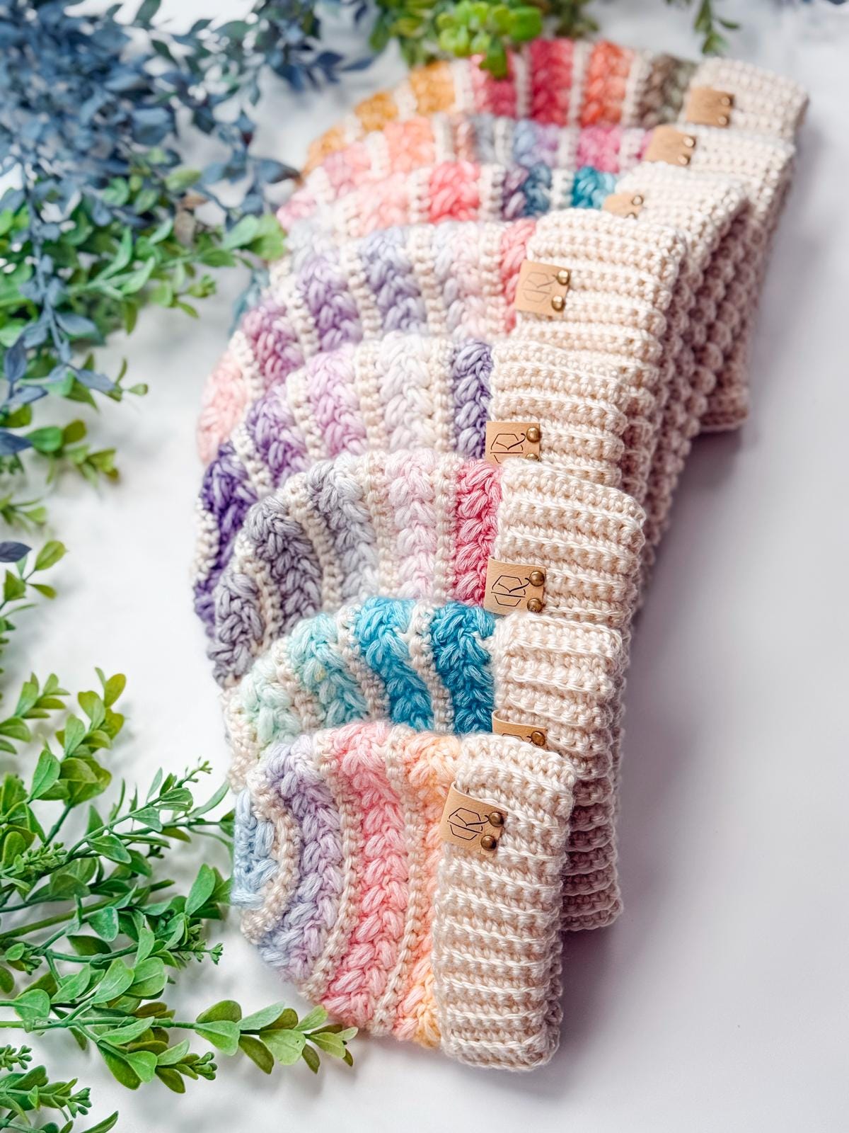 Seven colorful crocheted beanies with striped patterns are arranged on a white surface next to green leafy plants, making a charming handmade gift that pairs perfectly with vintage clothes.