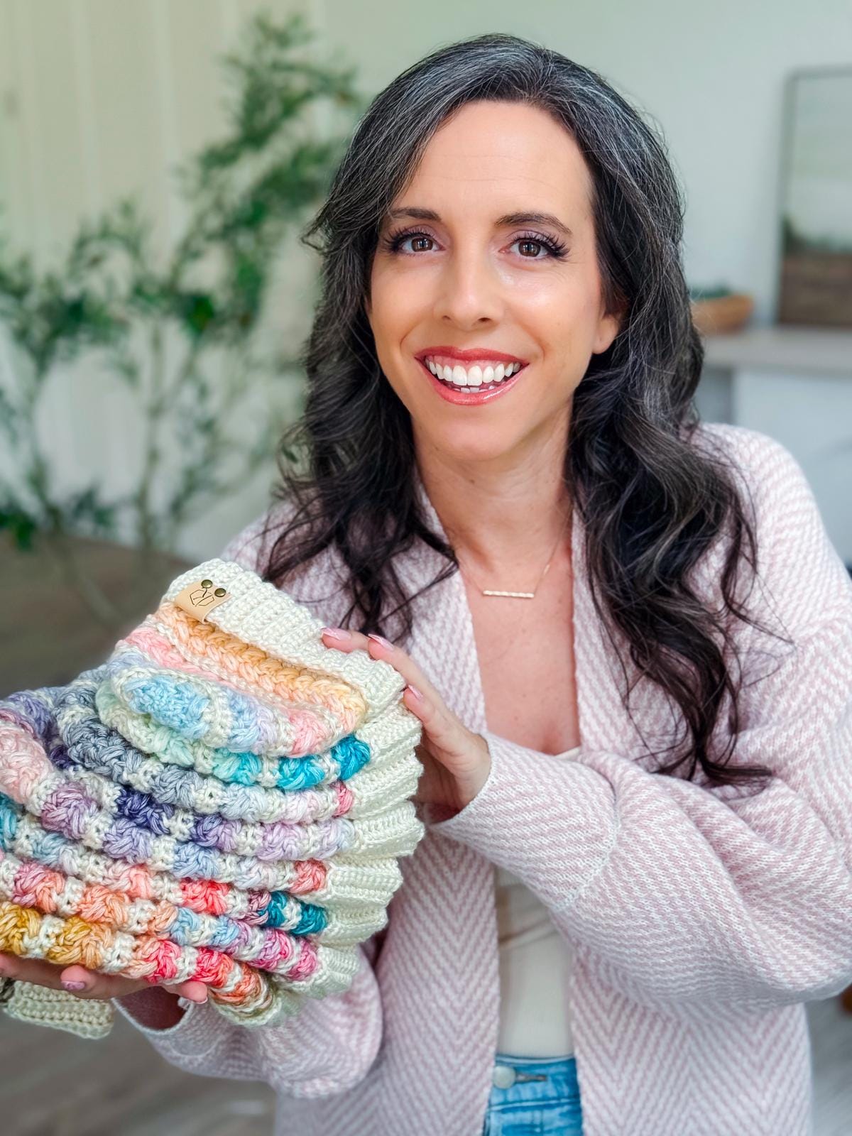 A woman with long dark hair and a light pink sweater holds a stack of colorful, crocheted handmade gifts and smiles at the camera indoors.