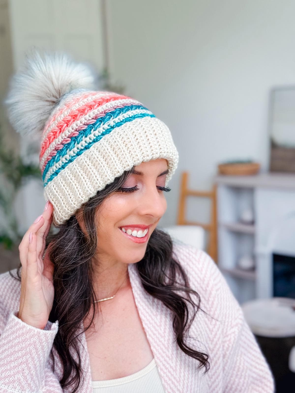 Woman wearing a crochet hat with colorful stripes and a pom-pom, smiling and looking down while sitting indoors, pairs her look with unique jewellery for a touch of charm.