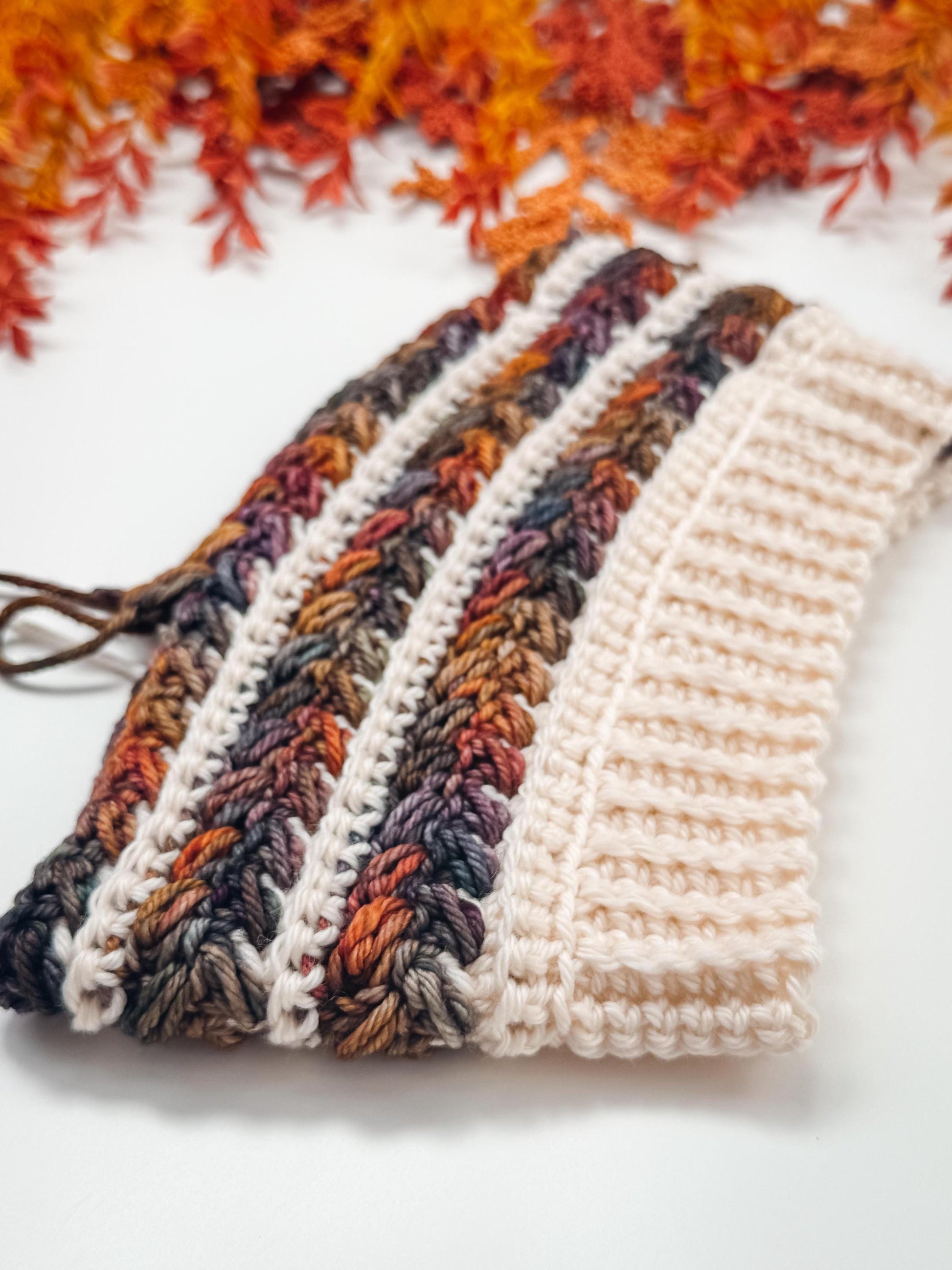A crocheted fabric with multicolored and cream yarn patterns is displayed on a white surface, with artificial autumn leaves in the background—perfect as a handmade gift or to complement your vintage clothes collection.