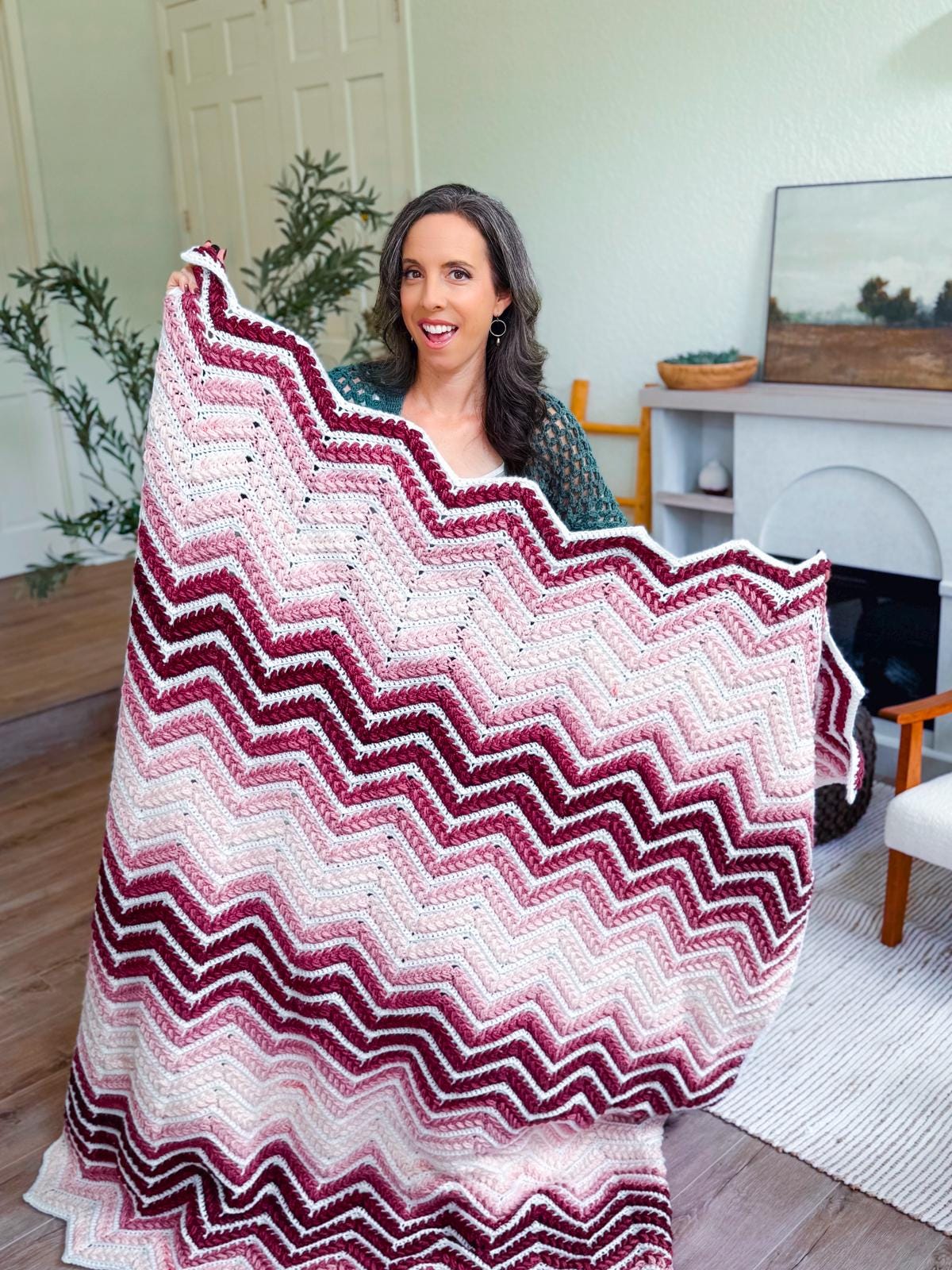 A woman sits indoors holding up a large zigzag-patterned crochet blanket in shades of burgundy, pink, and white—a cozy handmade gift or crochet home decor.