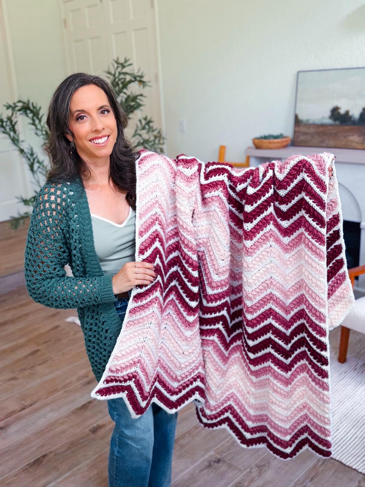 A woman stands indoors holding up a crocheted zigzag-patterned crochet blanket in shades of burgundy, pink, and white—a perfect handmade gift to cherish or share.