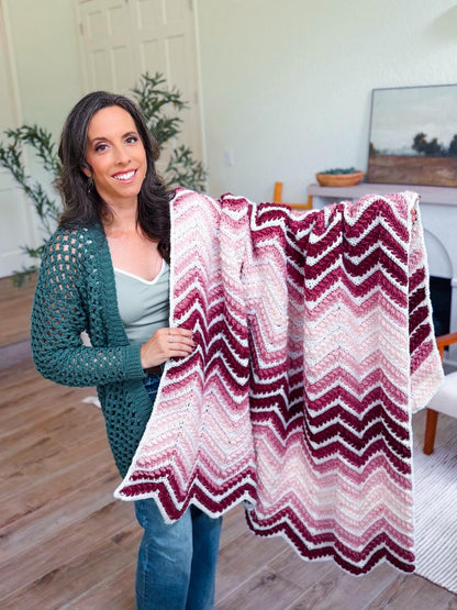 Pink, burgundy and white chevron crochet blanket held over the arm of a smiling woman in a green cardigan