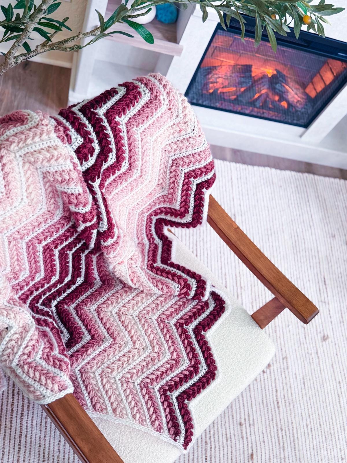 A pink and burgundy zigzag patterned knit blanket, perfect as a handmade gift, draped over a wooden chair beside a lit fireplace on a light striped rug.