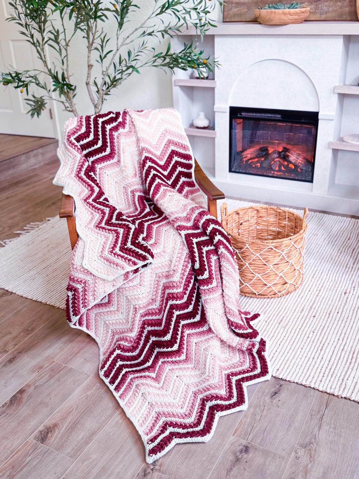 A zigzag-patterned crochet blanket in shades of pink, burgundy, and white draped over a wooden chair beside a wicker basket—perfect as a handmade gift—with a fireplace and houseplant in the background.