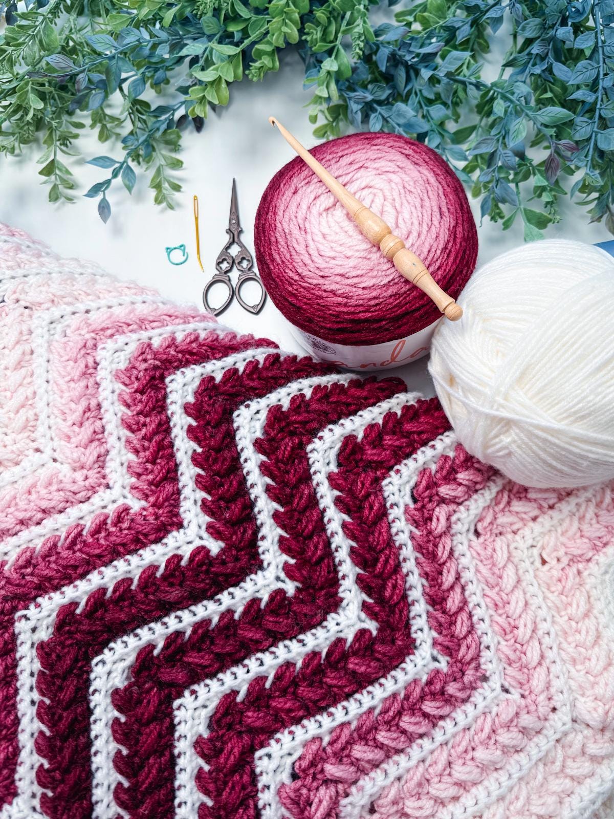 A chevron-patterned crochet blanket in shades of pink and white, yarn balls, crochet hooks, scissors, and greenery arranged on a white surface—perfect as a handmade gift or paired with unique jewellery for a thoughtful present.