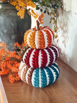 Stack of three striped crochet pumpkins in orange, red, and teal with white stripes on a wooden shelf