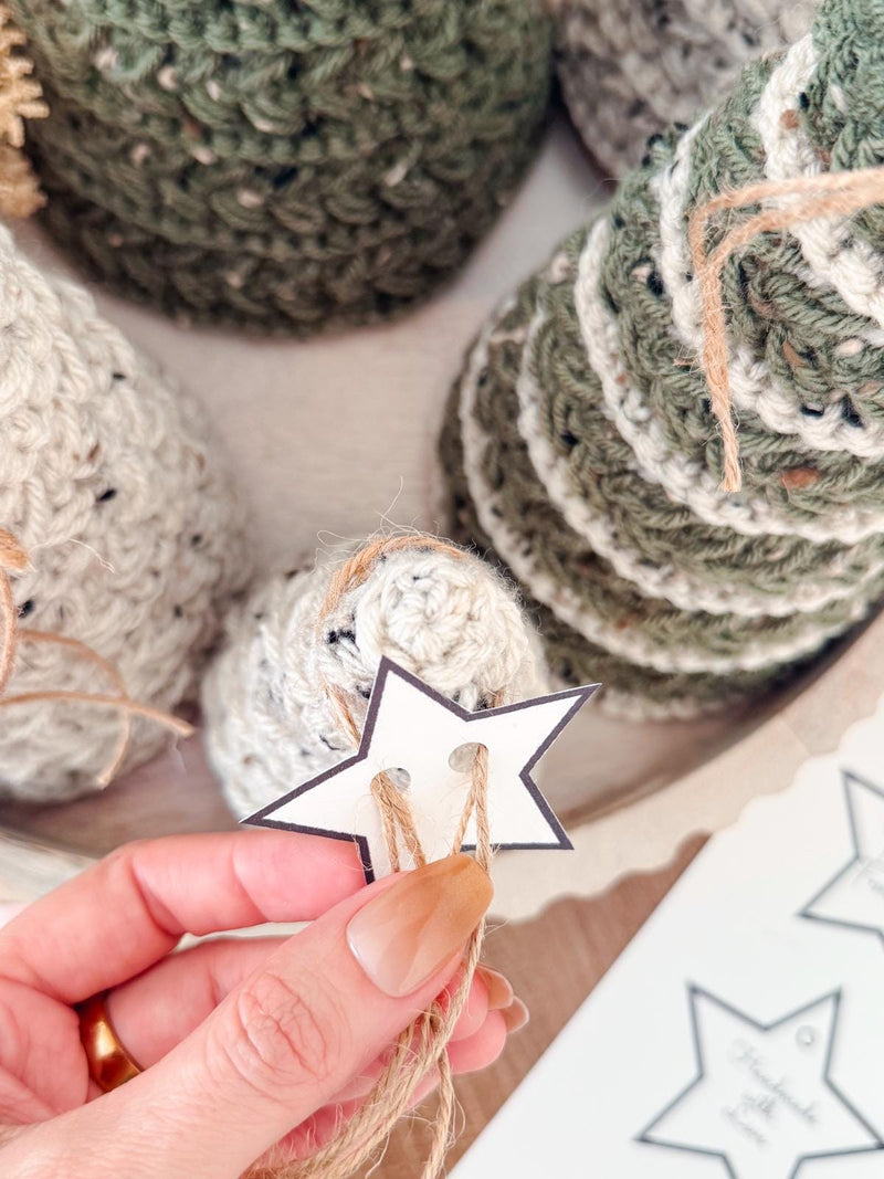 A hand holds a twine tag shaped like a star in front of crocheted cone-shaped decorations in green and beige tones, perfect as a handmade gift or to pair with unique jewellery for a charming display.